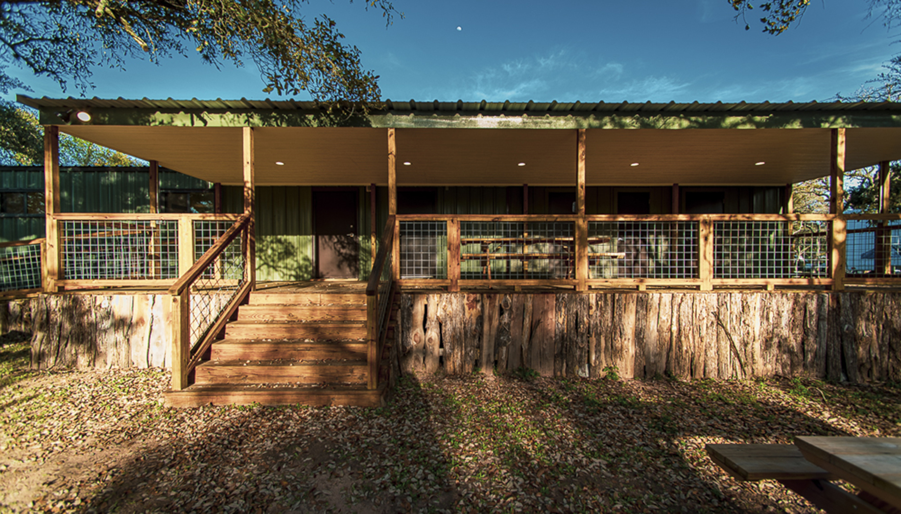 A green cabin with a metal roof and a wooden porch with stairs leading up to the entrance, surrounded by trees and fallen leaves on the ground.